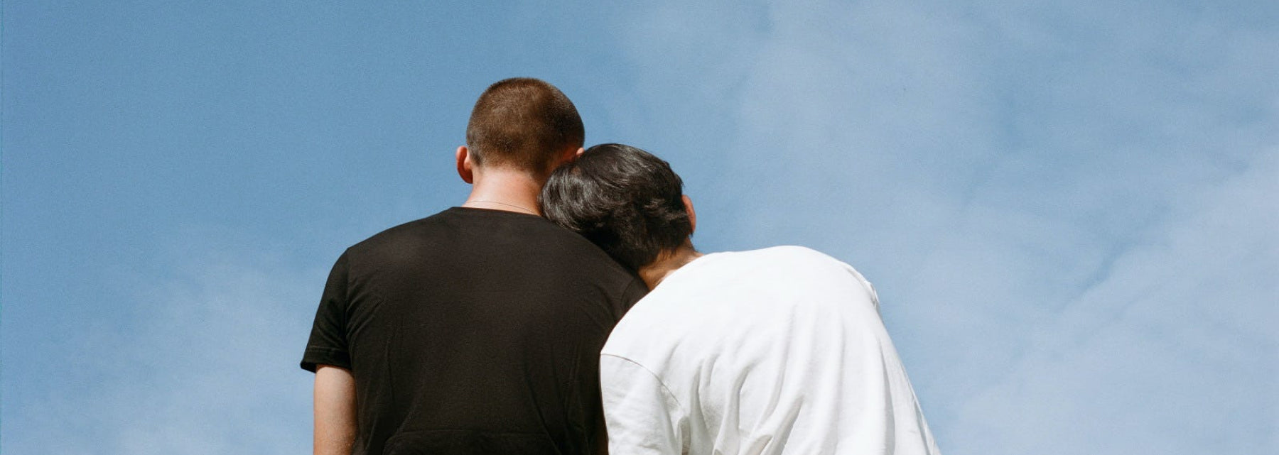 Two people lean against each other in front of a blue sky
